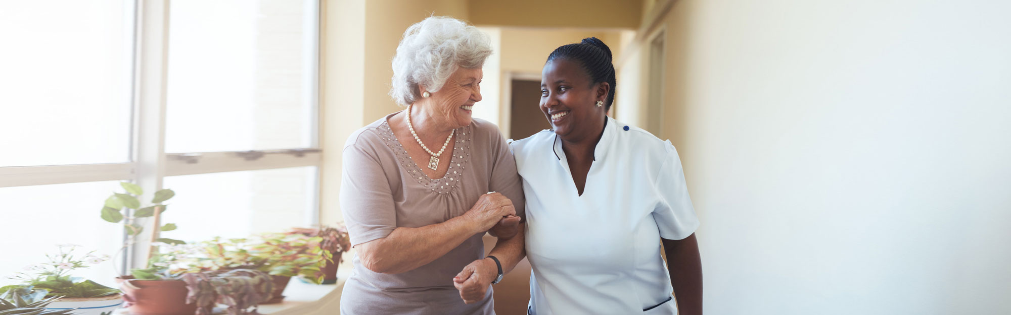 senior woman arm-in-arm with caregiver smiling while walking down a hallway